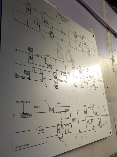 A large plastic engraved map showing the interior layouts of a few floors of Sumervale house, shown nailed to a slightly grubby/damaged lilac painted wall. The map has some graffiti scratched into it.