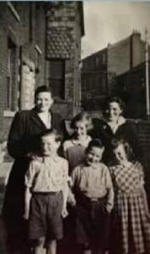 A black and white photo of a family of six, with two older women, two younger girls, and two younger boys. They’re standing on a street, with the view down the street behind them.